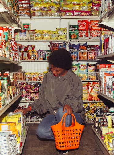 African American woman selecting products in a convenience store with an orange basket.
